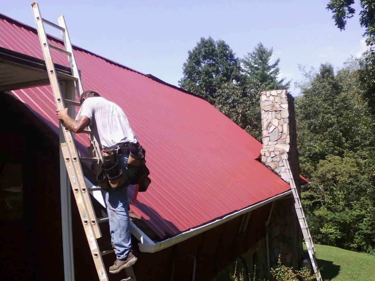 Roofer on a ladder beside a red metal roof and stone chimney — Soffit & Fascia Repair by Roofing Brothers in Cusick
